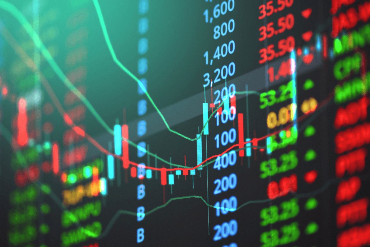 Online trading dashboard showing stock charts, candlestick graphs, live prices, and cryptocurrency icons on a laptop and smartphone in a modern workspace.